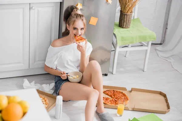 Young woman in shorts sitting on floor in kitchen and eating pizza and cornflakes — Stock Photo