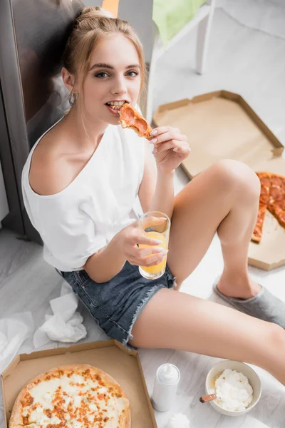 High angle view of pleased blonde woman eating pizza while sitting on floor in kitchen — Stock Photo