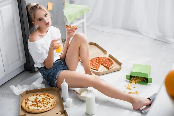Sensual young woman holding pizza and orange juice while sitting on floor in kitchen — Stock Photo