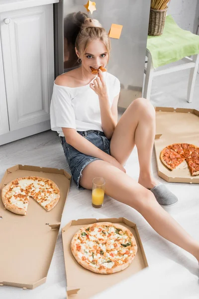 Seductive young woman sitting on floor in kitchen and eating pizza — Stock Photo