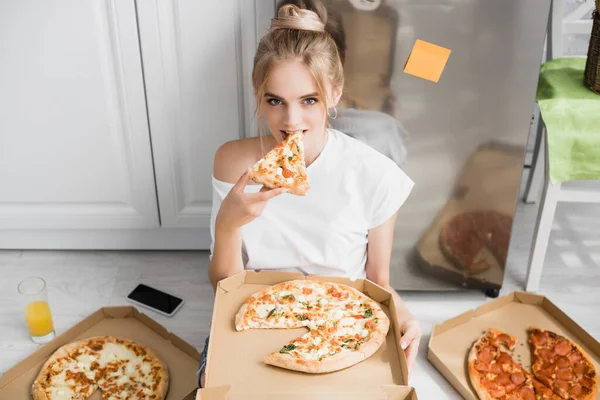 Junge blonde Frau blickt in die Kamera, während sie in der Küche auf dem Boden sitzt und Pizza isst — Stockfoto