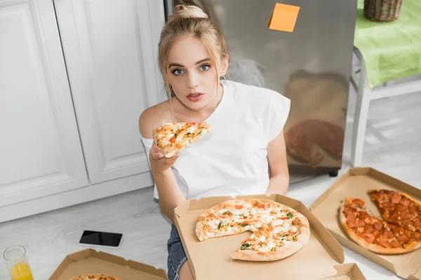 Visão de alto ângulo da jovem mulher sentada no chão na cozinha com pedaço de pizza — Fotografia de Stock