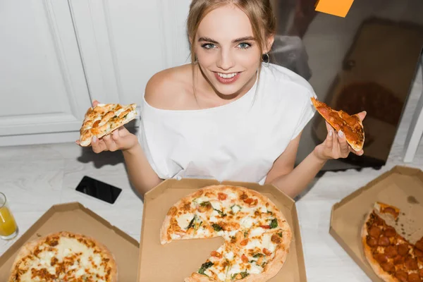 Vue grand angle de femme blonde joyeuse regardant la caméra tout en étant assis sur le sol dans la cuisine et tenant pizza — Photo de stock