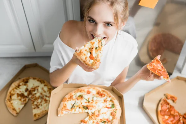 Vue grand angle de femme blonde manger de la pizza tout en étant assis sur le sol dans la cuisine — Photo de stock