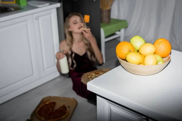 Focalizzazione selettiva della giovane donna che mangia pizza e tiene il latte seduto sul pavimento vicino al tavolo con frutta fresca — Foto stock