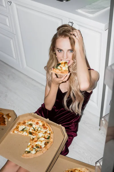 High angle view of sexy woman eating pizza and touching hair while sitting on floor and looking at camera — Stock Photo