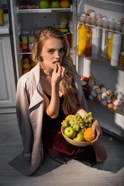 Sexy joven mujer en gabardina comer uva cerca abierto refrigerador - foto de stock