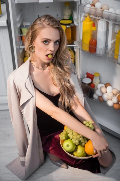 Sensual mujer joven comiendo uvas mientras sostiene frutas frescas cerca del refrigerador abierto - foto de stock