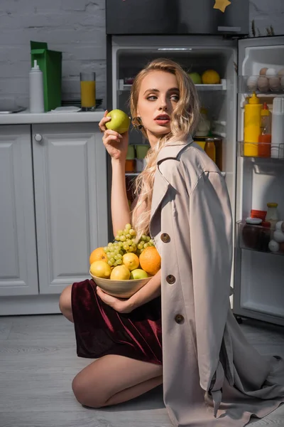 Donna bionda in trench cappotto contenente ciotola con frutta fresca vicino al frigorifero aperto — Foto stock