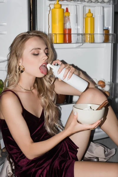 Сексуальна блондинка їсть збитих вершків з пляшки, сидячи біля холодильника з мискою — стокове фото