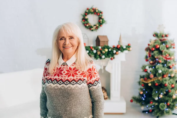 Joyeuse femme âgée souriant à la caméra près décoré arbre de Noël, branche et couronne sur fond — Photo de stock