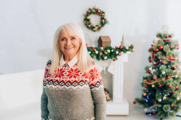 Mulher sênior alegre sorrindo para a câmera perto de árvore de natal decorada, ramo e grinalda no fundo — Fotografia de Stock