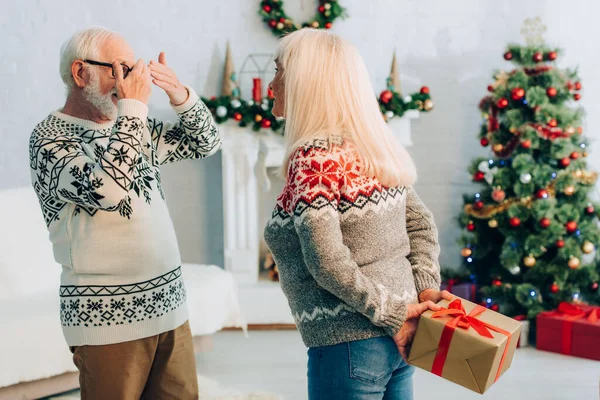 Senior Mann bedeckt Augen mit Händen, während Frau Geschenk hinter dem Rücken hält — Stockfoto