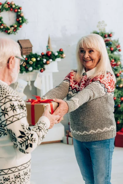 Избирательный фокус веселой пожилой женщины, преподносящей рождественский подарок мужу — стоковое фото