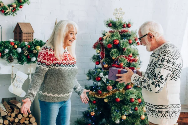 Senior man holding gift box near excited wife standing with open arms near christmas tree — Stock Photo