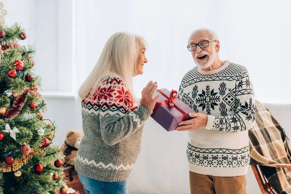 Hombre mayor emocionado presentando caja de regalo a marido feliz cerca del árbol de Navidad - foto de stock
