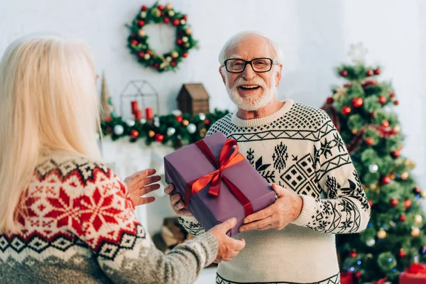 Selektiver Fokus eines glücklichen Seniors, der in die Kamera schaut, während er seiner Frau ein Weihnachtsgeschenk überreicht — Stockfoto