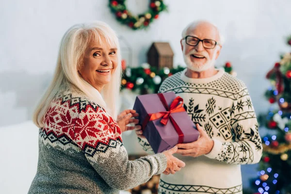 Fröhliche Seniorin blickt in die Kamera, während sie ihrem glücklichen Mann ein Weihnachtsgeschenk überreicht — Stockfoto