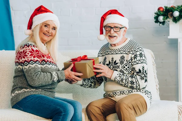 Felice coppia anziana in cappelli di Babbo Natale guardando la fotocamera mentre seduto sul divano con scatola regalo — Foto stock