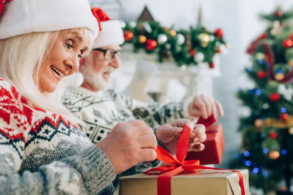 Selektiver Fokus der fröhlichen Seniorin beim Öffnen der Geschenkbox in der Nähe ihres Mannes — Stockfoto