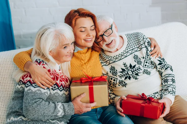 Happy woman with closed eyes embracing senior parents holding gift boxes — Stock Photo