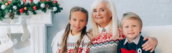 Plan panoramique de femme âgée gaie avec des petits-enfants regardant la caméra tout en étant assis près de la cheminée avec décoration de Noël — Photo de stock