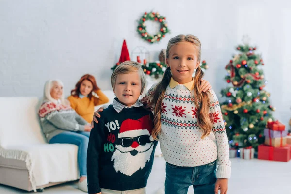 Glückliche Geschwister, die sich umarmen und in die Kamera schauen, während Mutter und Großmutter im Hintergrund sitzen — Stockfoto