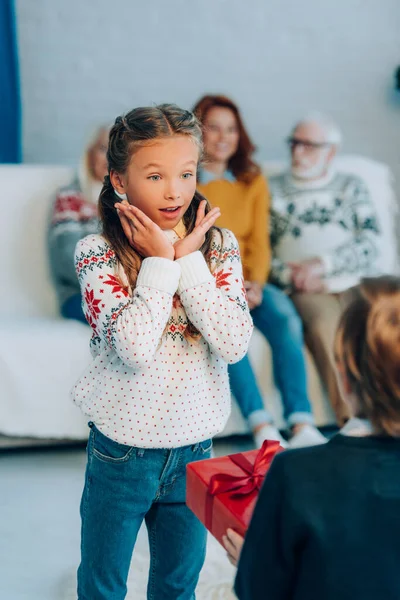 Foco seletivo da menina animado de mãos dadas perto do rosto enquanto olha para o irmão segurando presente de Natal — Fotografia de Stock