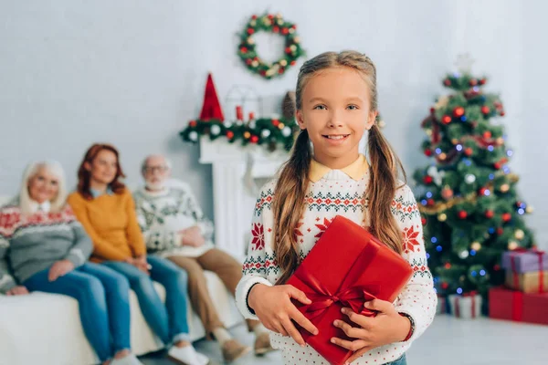 Menina feliz segurando caixa de presente e olhando para a câmera enquanto a mãe e os avós sentados no fundo — Fotografia de Stock