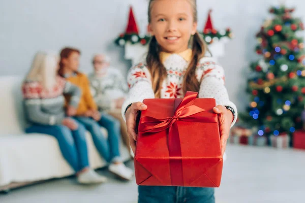 Fille souriante tenant boîte cadeau et regardant la caméra tandis que la famille assis sur le fond — Photo de stock