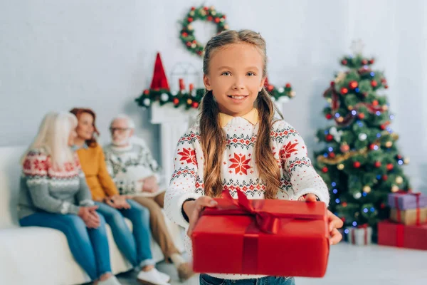Alegre chica sosteniendo navidad presente y mirando a la cámara mientras los familiares sentados en el fondo - foto de stock