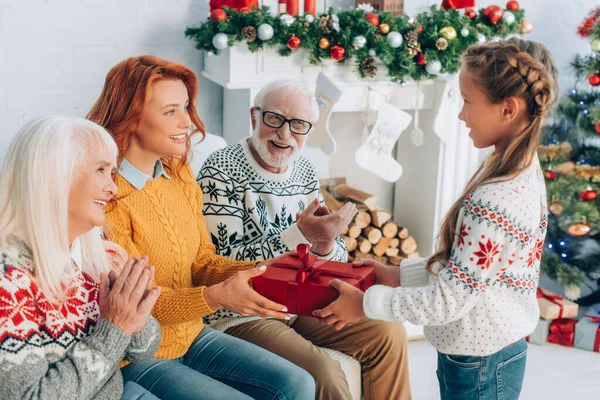 Lächelndes Mädchen überreicht Geschenkbox an glückliche Mutter in der Nähe applaudierender Großeltern — Stockfoto