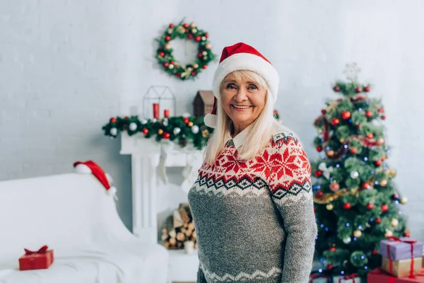 Joyeuse femme âgée en chapeau santa regardant la caméra près de la décoration de Noël sur fond — Photo de stock