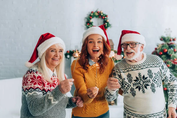 Excité couple aîné avec fille dans santa chapeaux tenant des étincelles sur Noël — Photo de stock