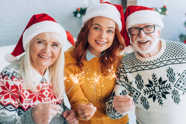 Joyful senior couple with daughter in santa hats holding sparklers while looking at camera — Stock Photo