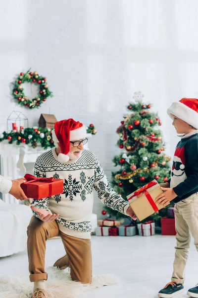 Großvater mit Weihnachtsmütze nimmt Geschenke von Enkeln an Weihnachten entgegen — Stockfoto