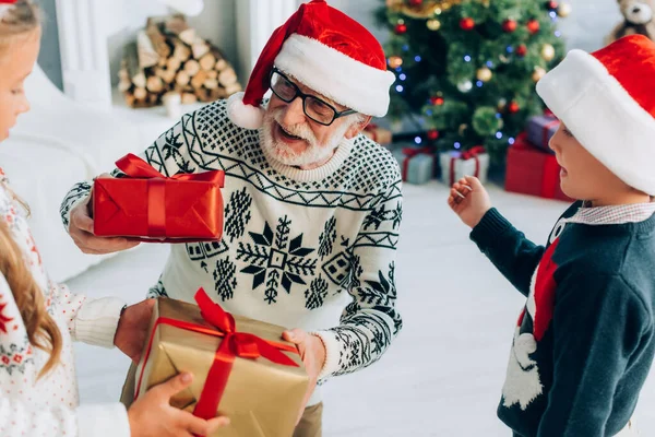 Girl and boy presenting gift boxes to happy grandfather in santa hat — Stock Photo