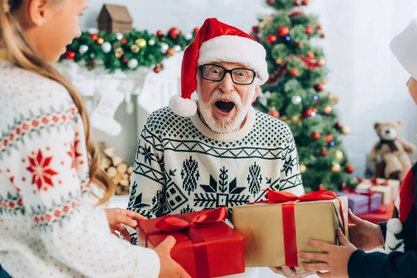 Aufgeregter Senior mit Weihnachtsmütze nimmt Weihnachtsgeschenke von Enkeln entgegen — Stockfoto