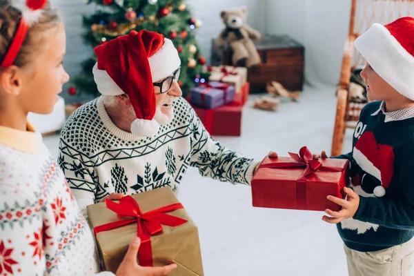 Senior mit Weihnachtsmütze nimmt Weihnachtsgeschenke von Enkeln entgegen — Stockfoto