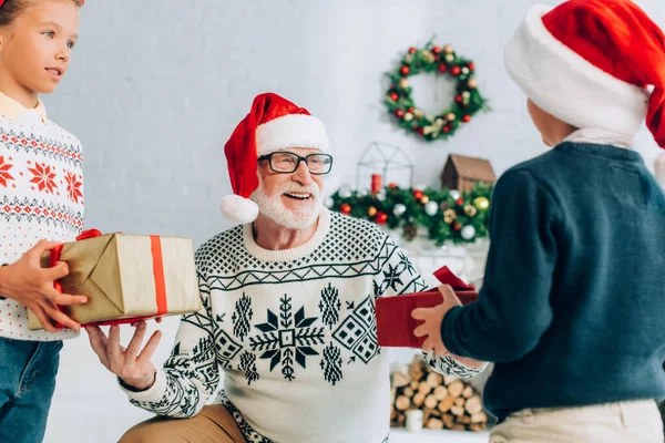Щасливий старший чоловік у капелюсі Санти бере різдвяні подарунки від онуків — стокове фото