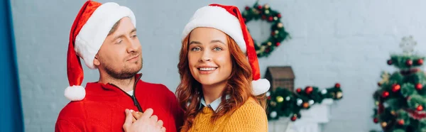 Horizontales Bild einer Frau mit Weihnachtsmütze, die in die Kamera lächelt, während sie ihren Mann an Weihnachten umarmt — Stockfoto