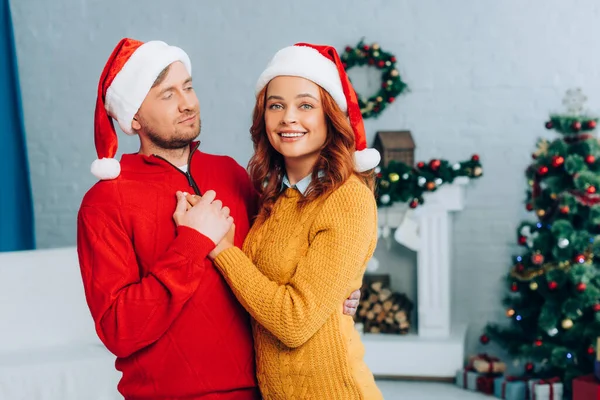 Glückliche Frau mit Weihnachtsmütze blickt in die Kamera, während sie ihren Mann an Weihnachten umarmt — Stockfoto