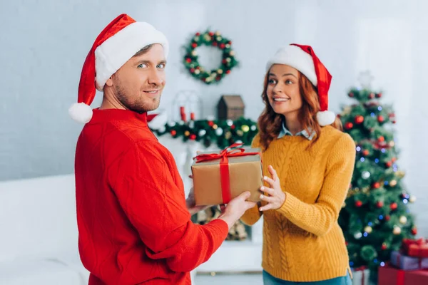 Lächelnder Mann mit Weihnachtsmütze blickt in die Kamera, während er seiner aufgeregten Frau eine Geschenkbox überreicht — Stockfoto