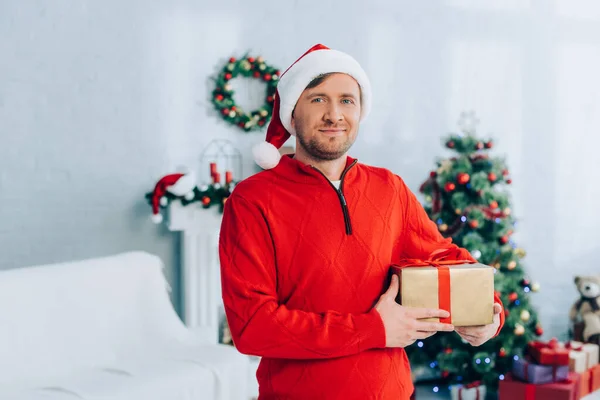 Homme en santa chapeau et pull regardant la caméra tout en tenant boîte cadeau — Photo de stock