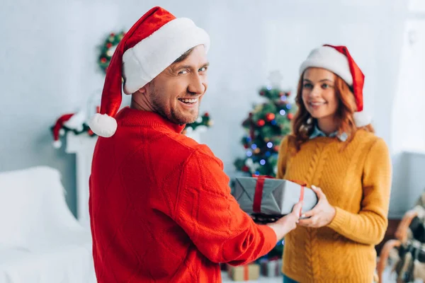 Fröhlicher Mann blickt in die Kamera, während er seiner lächelnden Frau eine Geschenkbox überreicht — Stockfoto