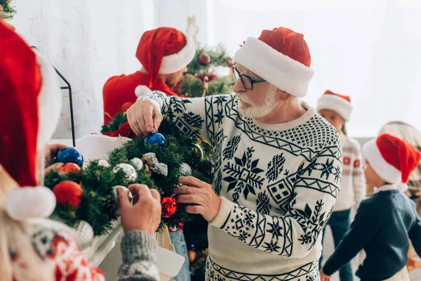 Senior man in santa hat decorating spruce branch with christmas baubles together with family — Stock Photo