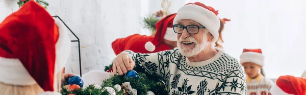 Panoramic shot of happy senior man in santa hat decorating pine branch together with family — Stock Photo