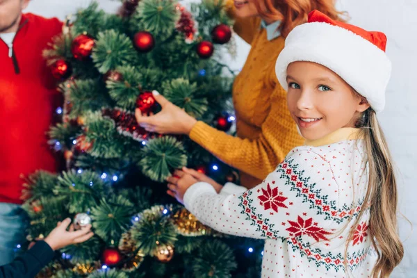 Glückliches Mädchen mit Weihnachtsmütze, das in die Kamera schaut, während es mit der Familie den Weihnachtsbaum schmückt — Stockfoto