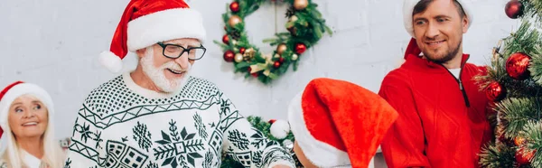 Concepto panorámico de hombre mayor en sombrero de santa mirando nieto cerca de la familia y el árbol de Navidad - foto de stock