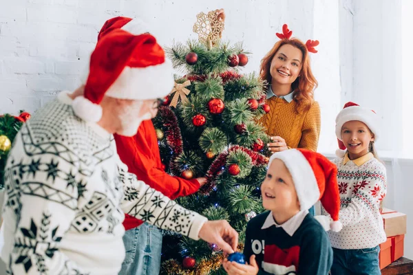 Selektiver Fokus des Großvaters, der dem Enkel eine Christbaumkugel schenkt, während er mit der Familie den Weihnachtsbaum schmückt — Stockfoto